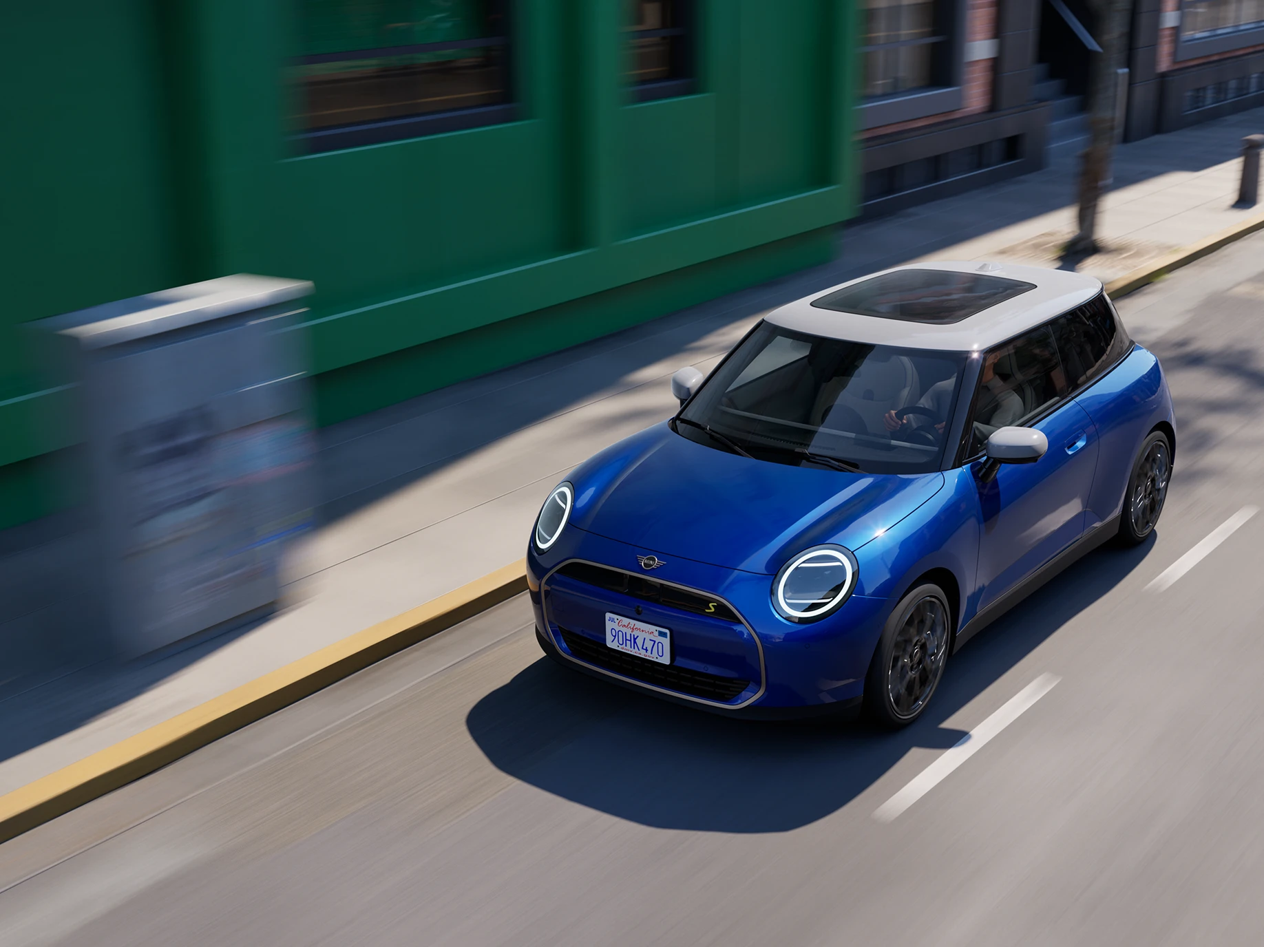Вигляд зверху повністю електричного MINI Cooper кольору Blazing Blue з білим дахом, що їде вулицею. 
