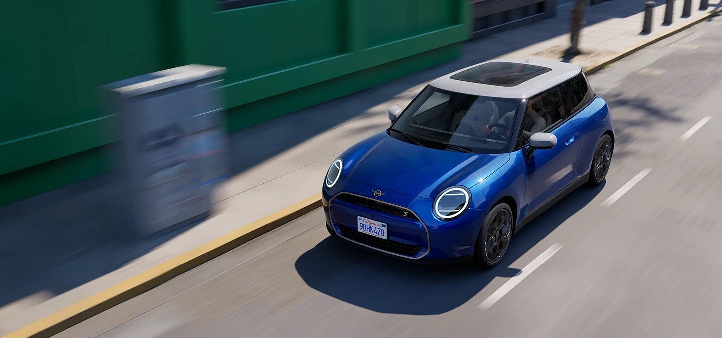 Вигляд зверху повністю електричного MINI Cooper кольору Blazing Blue з білим дахом, що їде вулицею. 