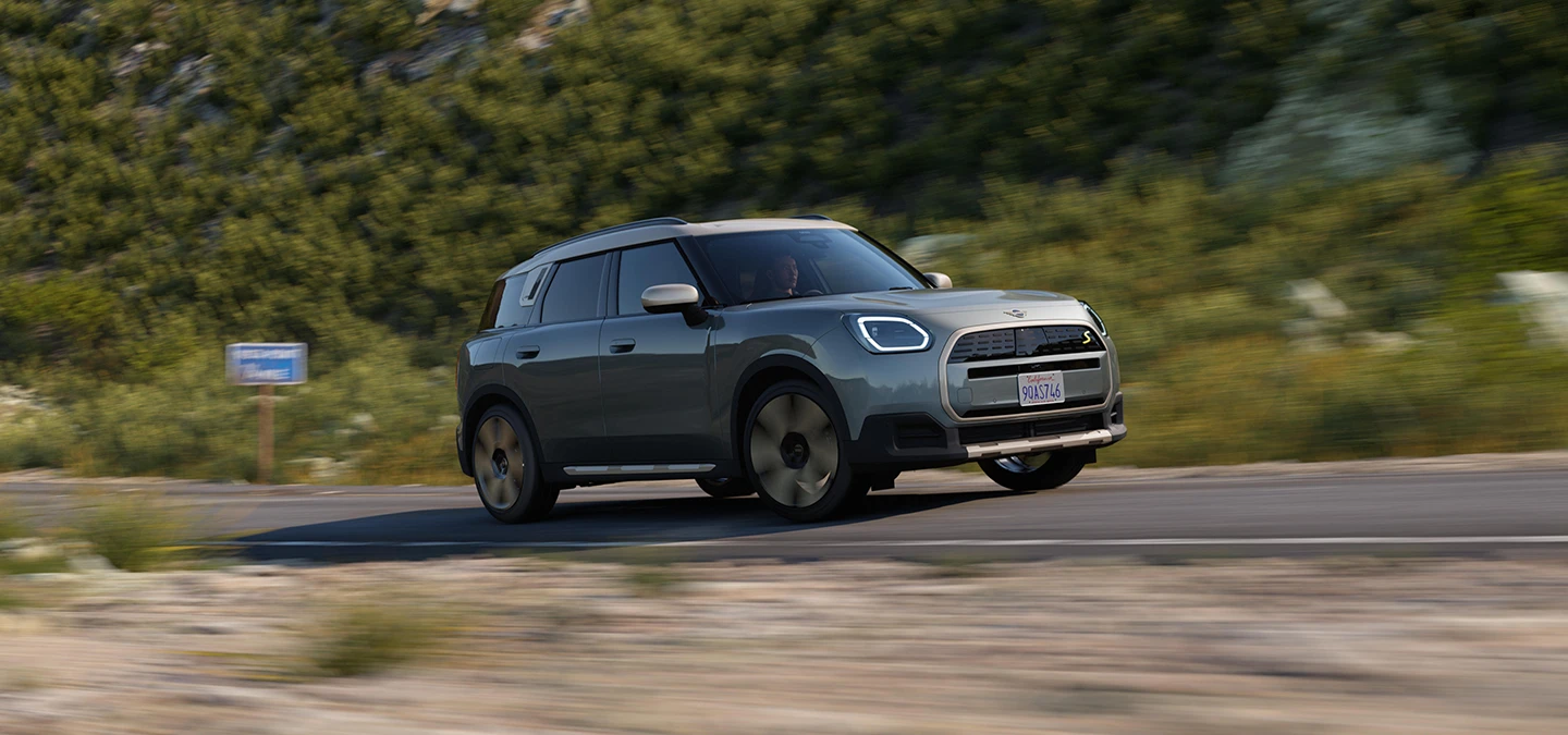Повністю електричний MINI Countryman в кольорі Smokey Green і з яскравим сріблястим дахом - вигляд збоку.